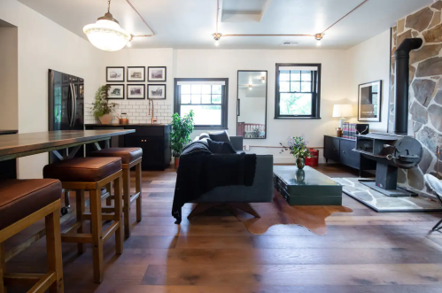 Cozy living room with a stone fireplace, modern furniture, and a dining area featuring bar stools and a large window.